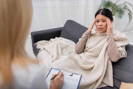 Asian woman holding napkin and suffering from headache near blurred doctor with clipboard at home 