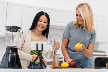 Smiling interracial bloggers holding water and banana near smartphone on tripod at home 