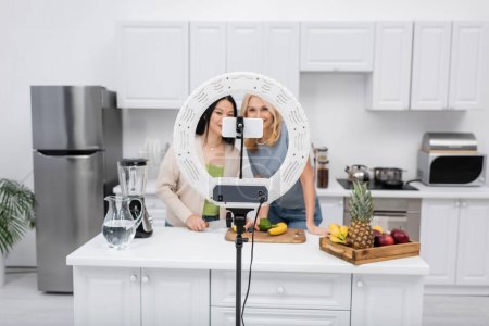 Foto de Lámpara de anillo y teléfono inteligente cerca de bloggers interracial borrosa y frutas frescas en la cocina - Imagen libre de derechos