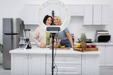Positive interrassische Blogger, die in der Nähe von Früchten in der Küche auf das Smartphone schauen 