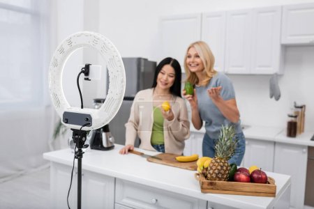 Foto de Lámpara de anillo y smartphone cerca de blogueros interracial borrosa con frutas maduras en casa - Imagen libre de derechos