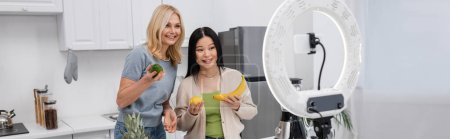 Interracial bloggers holding fresh fruits near cellphone in ring lamp at home, banner 