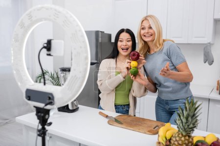 Téléchargez les photos : Blogueurs interraciaux excités tenant des fruits mûrs près du smartphone dans la cuisine - en image libre de droit