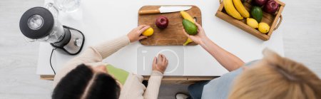 Photo for Overhead view of women holding ripe fruits near chopping board and blender in kitchen, banner - Royalty Free Image