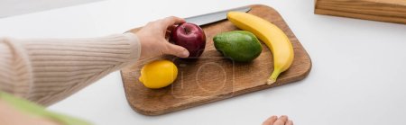 Cropped view of woman holding apple near fruits on chopping board at home, banner 