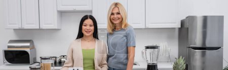 Positive multiethnic friends looking at camera near blender in kitchen, banner 