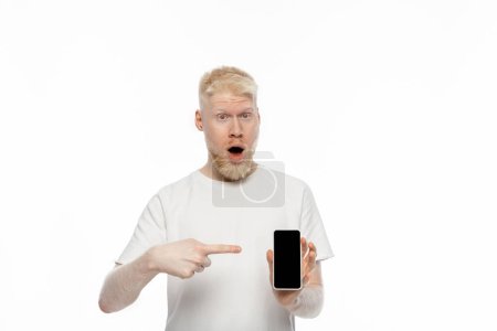 Foto de Impactado hombre albino en camiseta apuntando al teléfono inteligente con pantalla en blanco aislado en blanco - Imagen libre de derechos