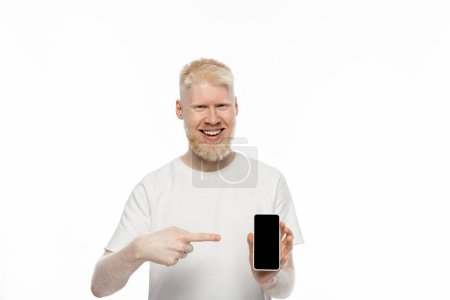 Foto de Hombre albino feliz en camiseta apuntando al teléfono inteligente con pantalla en blanco aislado en blanco - Imagen libre de derechos