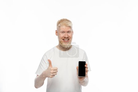 Foto de Hombre albino feliz en camiseta sosteniendo teléfono inteligente con pantalla en blanco y mostrando el pulgar hacia arriba aislado en blanco - Imagen libre de derechos