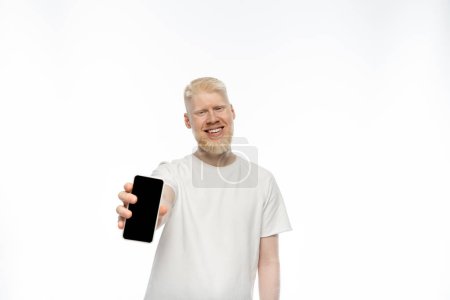 Foto de Hombre albino feliz en camiseta sosteniendo teléfono inteligente con pantalla en blanco sobre fondo blanco - Imagen libre de derechos
