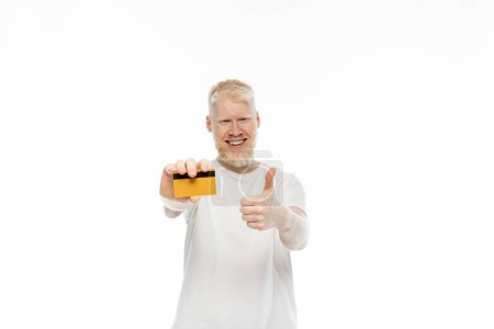 cheerful albino man in t-shirt holding credit card and showing thumb up isolated on white 
