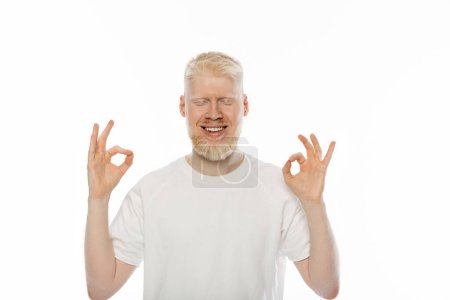 cheerful albino man with closed eyes showing ok sign with hands isolated on white 