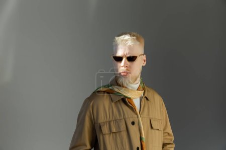 Sonnenlicht im Gesicht eines stilvollen Albino-Mannes mit Sonnenbrille und Hemdjacke auf grauem Hintergrund 
