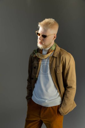 sunlight on face of stylish albino model in sunglasses posing with hands in pockets isolated on grey