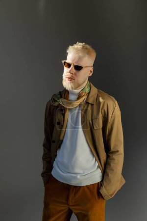 Sonnenlicht im Gesicht eines stilvollen Albino-Mannes mit Sonnenbrille, der mit Händen in Taschen auf grau posiert