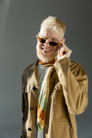 Sonnenlicht im Gesicht des glücklichen Albino-Mannes, der eine stylische Sonnenbrille aufsetzt und auf grau zwinkert 