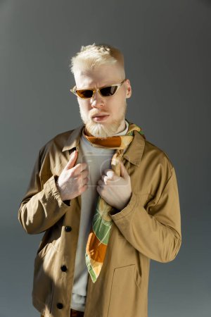 stylish albino man in sunglasses adjusting shirt jacket isolated on grey