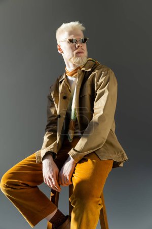 Téléchargez les photos : Élégant homme albinos en lunettes de soleil et veste de chemise assis sur chaise haute isolé sur gris - en image libre de droit