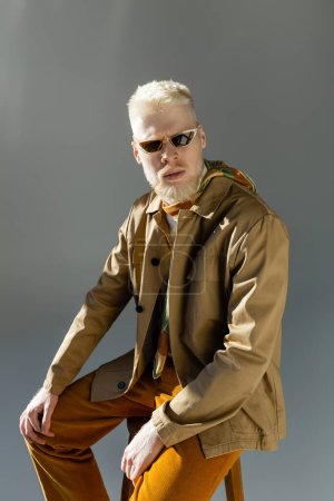 Téléchargez les photos : Homme albinos barbu dans des lunettes de soleil élégantes et veste de chemise assis sur chaise haute sur gris - en image libre de droit