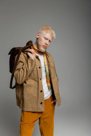 bearded albino man in stylish shirt jacket posing with hand in pocket and holding backpack isolated on grey
