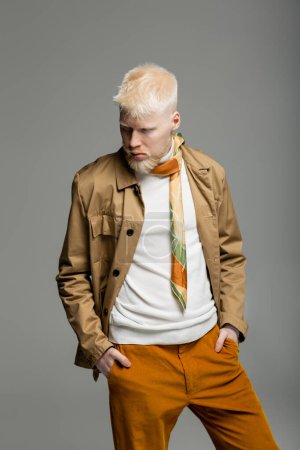 Albino-Mann in stylischer Hemdjacke und Seidenschal stehend, die Hände in Taschen isoliert auf grau 