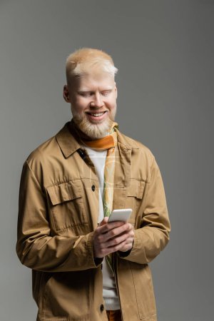 Foto de Hombre albino alegre en camisa elegante chaqueta y bufanda de seda de mensajería smartphone aislado en gris - Imagen libre de derechos