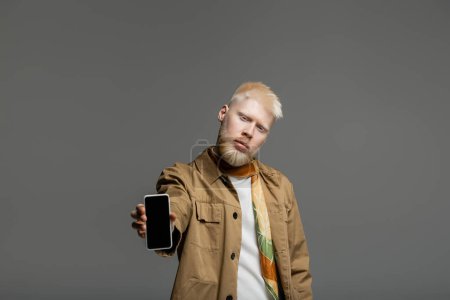 Foto de Hombre albino barbudo en chaqueta de camisa que sostiene el teléfono inteligente con pantalla en blanco aislado en gris - Imagen libre de derechos