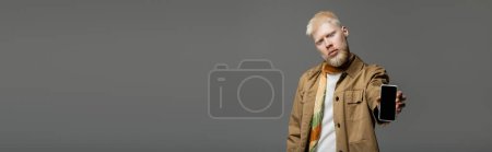 bearded albino man in shirt jacket holding smartphone with blank screen isolated on grey, banner 