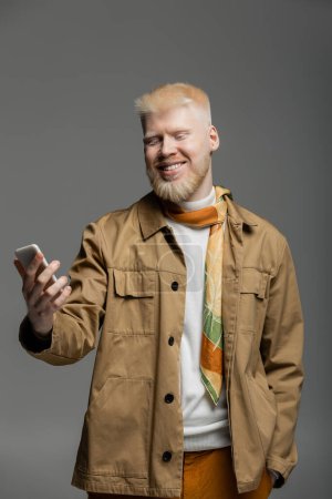 Foto de Hombre albino feliz en camisa chaqueta sosteniendo teléfono inteligente aislado en gris - Imagen libre de derechos
