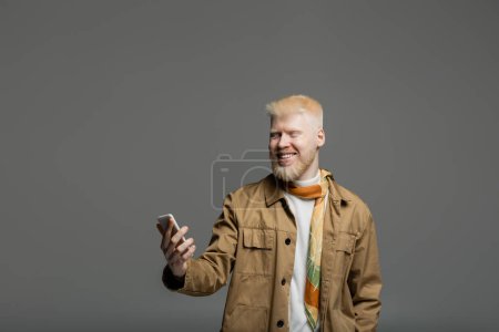 Téléchargez les photos : Homme barbu et heureux albinos en veste chemise en utilisant smartphone isolé sur gris - en image libre de droit
