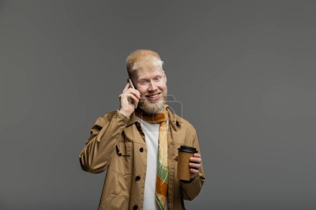 Foto de Hombre albino feliz en camisa chaqueta sosteniendo taza de papel y hablando en el teléfono inteligente aislado en gris - Imagen libre de derechos