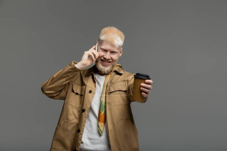 smiling albino man in shirt jacket holding paper cup and talking on smartphone isolated on grey