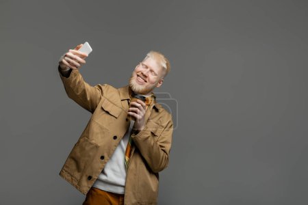 pleased albino man in shirt jacket holding paper cup and taking selfie on smartphone isolated on grey