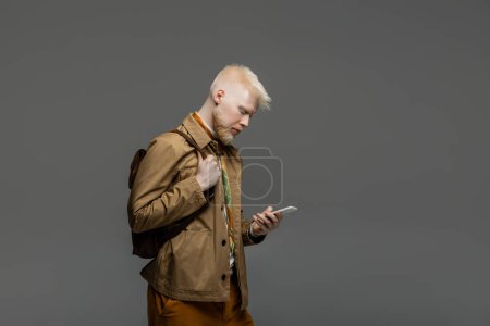 Foto de Hombre albino barbudo de pie con mochila y el uso de teléfono inteligente aislado en gris - Imagen libre de derechos