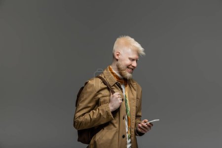 Foto de Hombre albino alegre de pie con mochila y el uso de teléfono inteligente aislado en gris - Imagen libre de derechos
