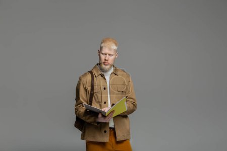 bearded albino student with backpack holding notebook isolated on grey