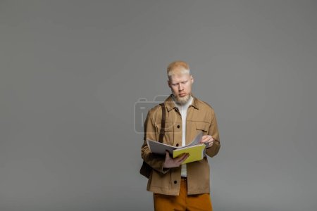 smart albino student with backpack holding notebook isolated on grey