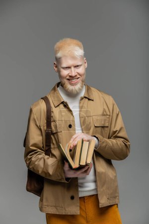 fröhlicher Albino-Student mit Rucksack, der Bücher isoliert auf grau hält