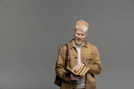 estudiante albino complacido con mochila sosteniendo libros aislados en gris