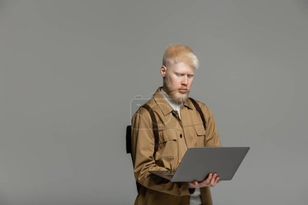 albino student with backpack using laptop while studying online isolated on grey