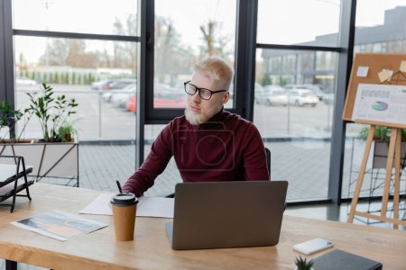 bärtiger Albino-Geschäftsmann mit Brille in der Nähe von Gadgets und Pappbecher auf dem Schreibtisch