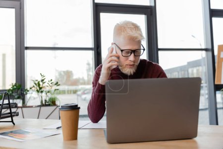 bearded albino businessman in glasses talking on smartphone near laptop and paper cup on desk