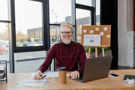 cheerful albino businessman in glasses looking at laptop near paper cup and infographics on desk  