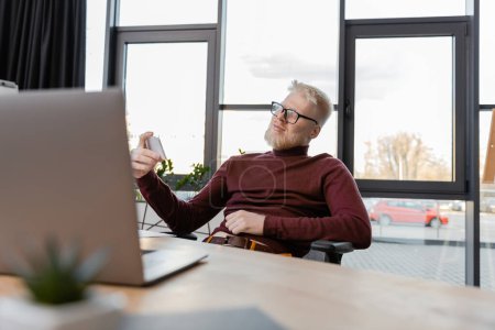 Albino-Geschäftsmann mit Brille, Smartphone und lächelndem Laptop im Büro 