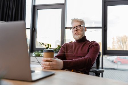 bearded albino businessman in glasses holding paper cup and looking at laptop on desk 