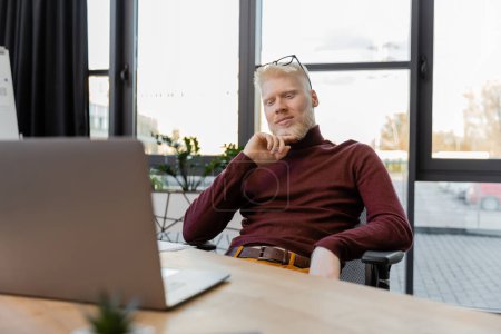 Nachdenklicher Albino-Geschäftsmann mit Brille blickt auf Laptop auf Schreibtisch 