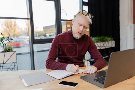 Bärtiger Albino-Geschäftsmann mit Brille schreibt auf Notizbuch neben Gadgets auf Schreibtisch 