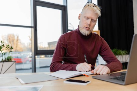 bearded albino businessman writing on notebook near devices on desk 