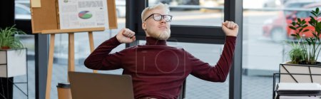 bearded albino businessman in glasses stretching near laptop in office, banner