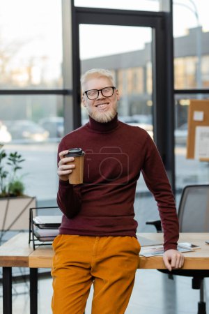 bearded albino businessman in glasses smiling and holding coffee to go in office